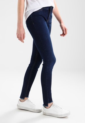 Donkerblauwe skinny jeans met een vervaagde zoom, voorzien van gouden stiksels en een mid-rise taille, gecombineerd met witte sneakers.