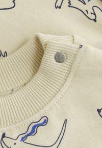 Cremfarbener gerippter Strickkragen eines Babybekleidungsstücks mit einem metallischen Druckknopf, der ein vierblättriges Kleeblatt zeigt, sowie einfachen blauen Tierumrissen auf dem Stoff.