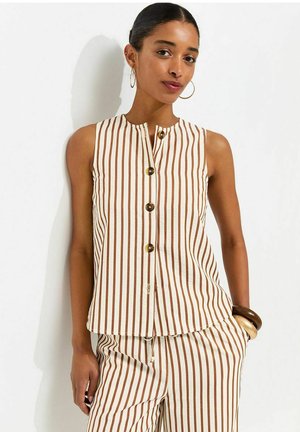 Donna che indossa un top bianco senza maniche e pantaloni con righe verticali marroni, grandi orecchini a cerchio e bracciali spessi.