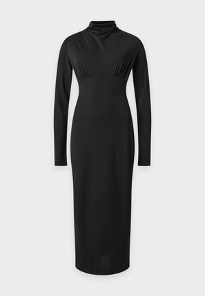 Une femme porte une robe midi noire à manches longues, un sac à main noir et des chaussures à talons à brides et à motifs, sur fond de studio blanc.