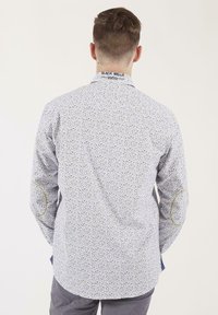 Chemise blanche avec un petit motif floral multicolore, manches longues, ourlet arrondi et coutures d' accent jaunes sur les coudières.