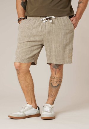 Homme portant un short en lin beige avec cordon de serrage, des baskets blanches et des tatouages sur les jambes, debout les mains dans les poches.