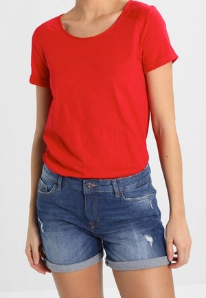 Woman wearing a bright red short-sleeve shirt and blue rolled-hem denim shorts with slight distressed details.