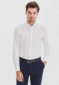Biała koszula z długim rękawem i kołnierzykiem na guziki. Gładka bawełniana tkanina, dopasowany krój, z kieszenią na lewej piersi. W zestawie z ciemnymi spodniami.