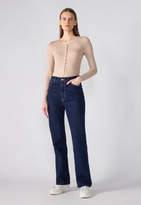 Bodysuit beige à manches longues avec boutons sur le devant, associé à un jean taille haute bleu foncé avec poches latérales et coutures contrastées. Baskets blanches.