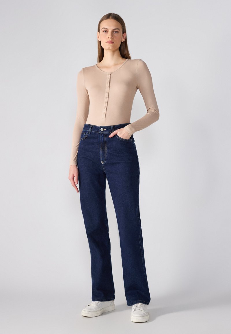 Bodysuit beige à manches longues avec boutons sur le devant, associé à un jean taille haute bleu foncé avec poches latérales et coutures contrastées. Baskets blanches.