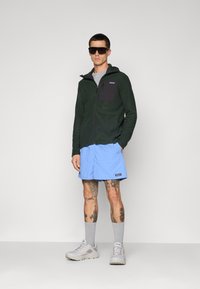 Vīrietis, tērpies tumši zaļā ar rūtiņu zippers, gaiši zilos šortos, pelēkās zeķēs, baltos sporta apavos un lielās melnās saules brillēs, stāv uz balta fona.