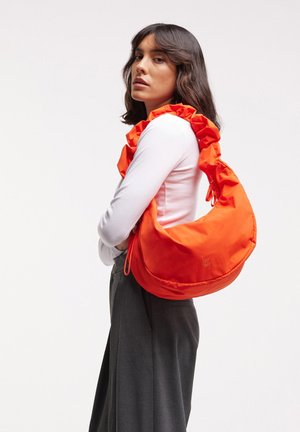 Vrouw met donker haar draagt een wit shirt, grijze broek en heeft een grote feloranje gefronste schoudertas bij zich.