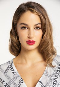 Blondes Haar mit sanften Wellen, trägt eine weiße Bluse mit schwarzem Kettenmuster und kräftigem roten Lippenstift, der einen klaren Teint betont.