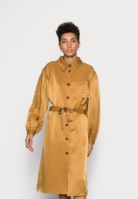 Vestido abotoado, de mangas longas, numa rica cor mostarda. Apresenta um colarinho, bolso frontal e um cinto na cintura. Tecido suave e brilhante com textura lisa.