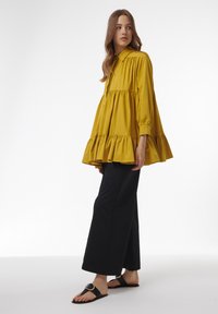 Blusa amarilla con cuello, volantes en capas, mangas largas, combinada con pantalones de pierna ancha negros y sandalias negras con una hebilla circular.