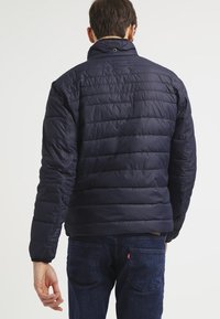 Uomo che indossa un giubbotto trapuntato blu navy e jeans blu scuro, girato di spalle contro uno sfondo bianco uniforme.