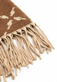 Brązowy materiał w rogu z kremowymi motywami ptaków i beżowymi skręconymi frędzlami wiszącymi na białym tle.