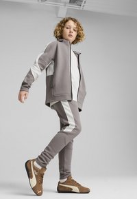 Grå atletisk tracksuit med hvide detaljer, lavet af blødt stof; har en lynlåsjakke og tætsiddende bukser. Brune træningssko inkluderet.