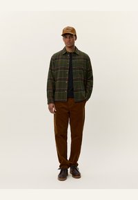 Groen geruite overhemden met een zwarte T-shirt, bruine ribfluwelen broek en bruine schoenen. Een beige pet met logo aan de voorkant accentueert de outfit.