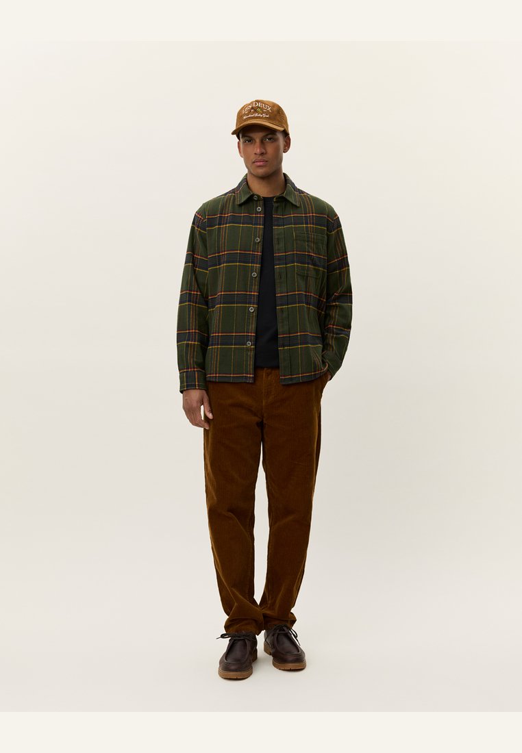 Groen geruite overhemden met een zwarte T-shirt, bruine ribfluwelen broek en bruine schoenen. Een beige pet met logo aan de voorkant accentueert de outfit.