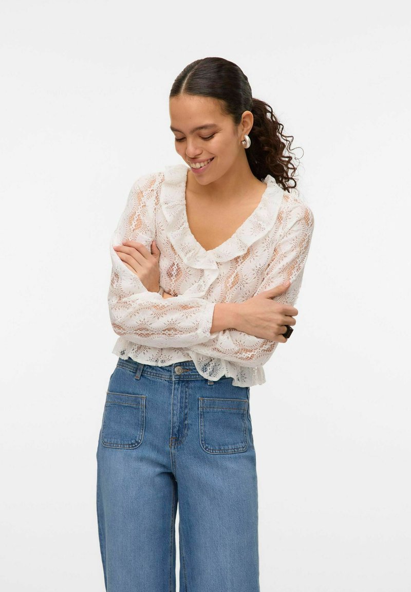 Blouse en dentelle blanche avec un col froncé et des manches longues, associée à un jean en denim bleu taille haute avec des poches avant.