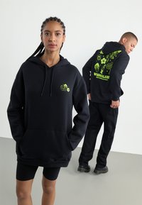 Black oversized hoodie featuring a front pocket and a neon green graphic design on the chest and back. Material: soft, textured fabric.