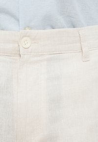 Μπεζ λινό παντελόνι με υφασμάτινη υφή, με κλείσιμο με ένα κουμπί στη μέση και λεία επιφάνεια.