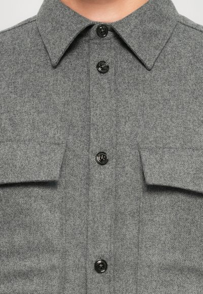 Gray wool shirt with a button-down collar, black buttons, and two chest pockets. Features a soft, textured fabric with a uniform design.