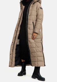 Mujer vestida con un abrigo largo acolchado beige sobre un vestido negro de canalé y botas negras con suela gruesa, de pie con el abrigo abierto.