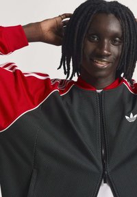 Čierna a červená športová bunda na zips s textúrou z mriežky, bielymi pruhmi a malým bielym logom na ľavej strane hrudníka. Dizajn s priliehavým golierom.
