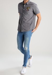Grijze spijkerblouse met korte mouwen met een geborduurd tijgerpatch, gecombineerd met lichtblauwe distressed skinny jeans en witte sneakers.