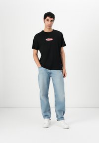 Black cotton t-shirt with red "GUESS JEANS" logo, paired with light blue distressed denim jeans and white sneakers. Model stands against a white background.