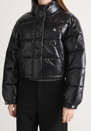 Žena v černé krátké lesklé péřové bundě s vysokým límcem a logem Calvin Klein na hrudi, kombinované s černými kalhotami.