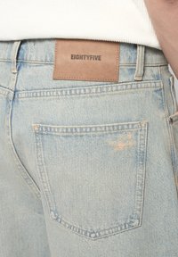Los jeans de mezclilla azul claro cuentan con un bolsillo trasero oculto y un parche de cuero marrón etiquetado como "EIGHTYFIVE" encima de la cinturilla. Textura descolorida y detalles de costura.