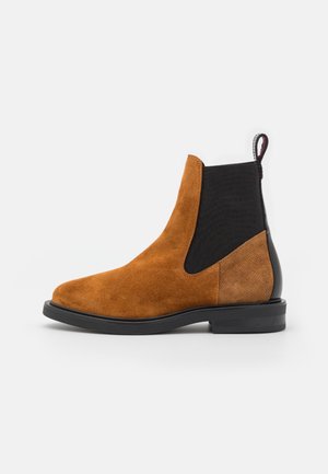 Bottine Chelsea en daim marron avec panneau élastique noir sur le côté, talon arrière texturé, languette de tirage et semelle noire sur fond blanc.