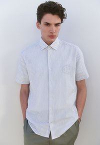 Chemise à manches courtes boutonnée avec rayures verticales bleu clair et blanc. Dotée d'un col italien et d'un tissu lisse et texturé.