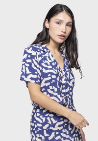 Blusa a maniche corte, con bottoni, di colore blu con un motivo astratto crema. Presenta un collo a V e una vestibilità rilassata. Tessuto liscio, dalla consistenza morbida.