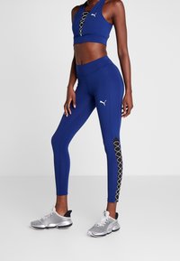 Modrý sportovní crop top s šněrováním v přední části a vysokými legínami s výřezem a černým šněrováním. Stříbrné tenisky.