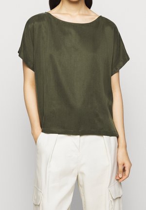 Kvinde iført olivengrøn kortærmet top med bådformet hals og hvide cargobukser med lommer, stående med den ene hånd i lommen.