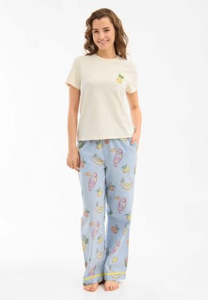 Młoda kobieta uśmiechająca się, w beżowej koszulce z małym wzorem ananasa i jasnoniebieskich spodniach do piżamy z kolorowymi nadrukami owoców i ptaków.