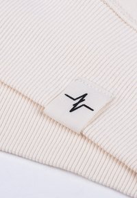 Rippiger cremefarbener Stoff mit einem gewebten Etikett, das ein schwarzes Blitzlogo zeigt. Glatte Textur, weicher und leichter Eindruck.