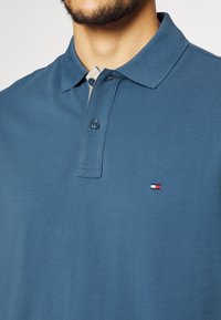 Mies, který nosí modrou polo košili se zapnutými dvěma knoflíky a malým červeným, bílým a modrým obdélníkovým logem na prsou.