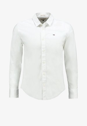 Biała koszula na guziki z kołnierzykiem, noszona z czarnymi spodniami i czarnymi skórzanymi butami. Koszula ma małe logo na piersi.