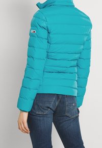 Tyrkysová bundu typu puffer s vysokým límcem, prošívaným designem a logem na rukávu, v kombinaci s tmavými džínami.