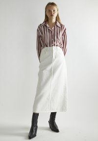 Camisa de botón a rayas en rojo y blanco, combinada con una falda maxi de mezclilla blanca. La modelo lleva botas de tobillo negras con tacón y detalles de tachuelas.