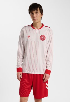 Jonge man draagt een wit voetbalshirt met lange mouwen met rode fijne strepen en rode korte broek, staand tegen een effen achtergrond.