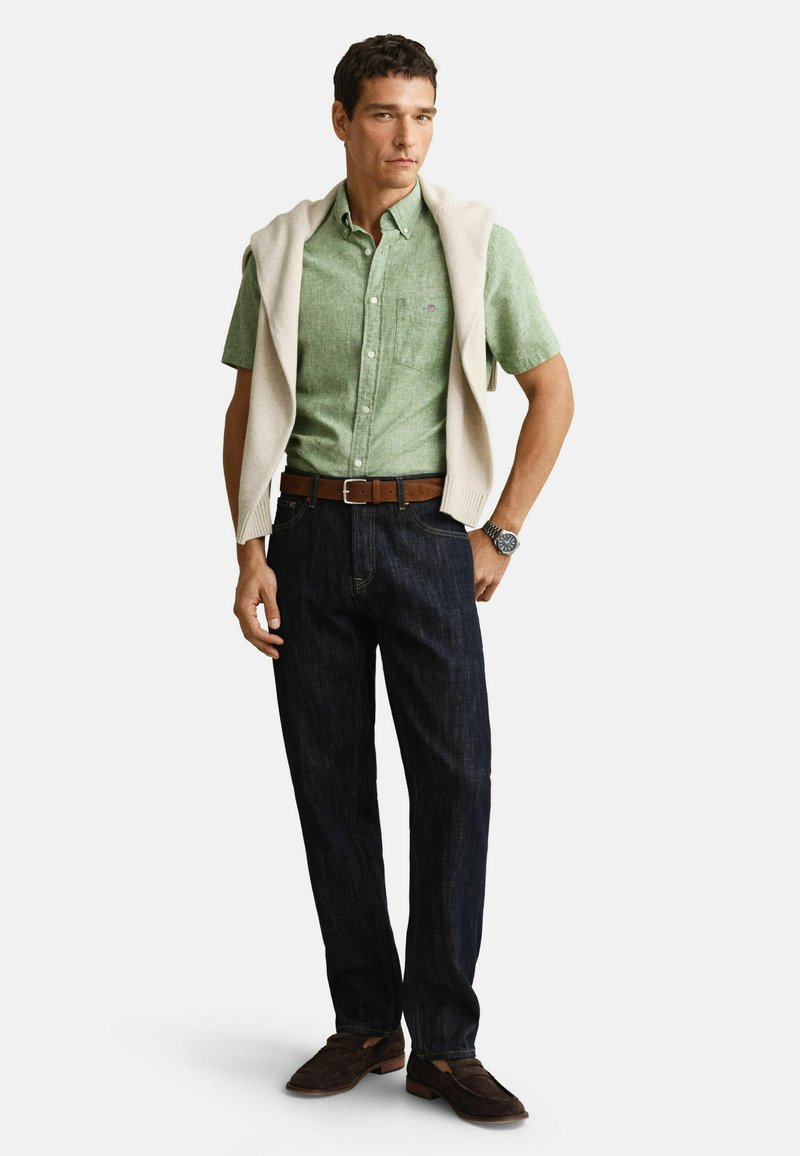 Mand iført en lysegrøn kortærmet skjorte med knapper, beige sweater lagt over skuldrene, mørke jeans, brun bælte, ur og brune sko.