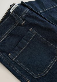 Džíny s širokými nohavicami z tmavozeleného denimového materiálu, s kontrastným žltým prešívaním, zadnou vreckou a štítkom s nápisom "WIDE LEG" na páse.