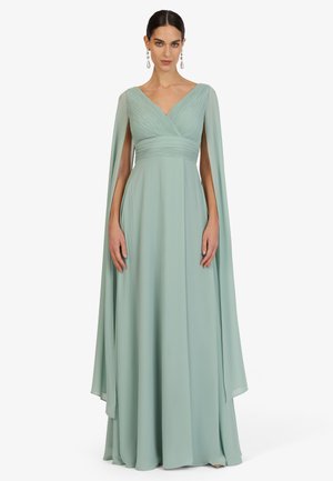 Woman wearing a floor-length light green gown with pleated V-neck bodice and attached flowing cape sleeves, standing against a plain background.