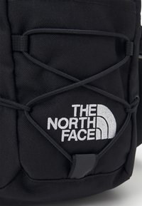 Zaino in tessuto nero con un gancio in gomma e logo ricamato bianco. Presenta un design con corda bungee per una maggiore flessibilità di stoccaggio.