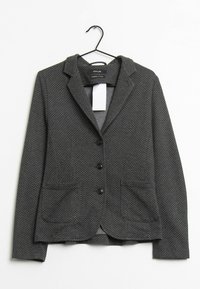 Blazer noir à pois blancs, design à simple boutonnage, revers crantés, deux poches avant et boutons détaillés, suspendu à un crochet noir.
