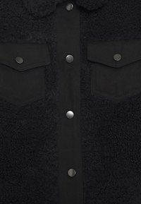 Black jacket featuring a textured fleece body, denim front, snap buttons, and two chest pockets with flap closures.
