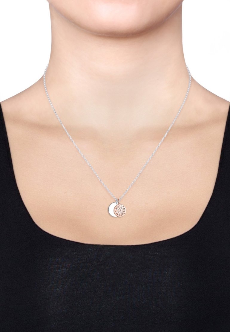 Sølv kædehalssmykke med to vedhængsdisker: en ensfarvet og en med indviklet mønster, der viser en blanding af metaller og teksturer.