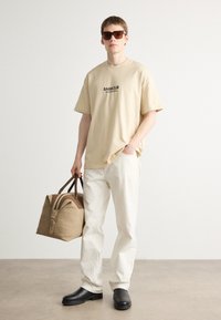 Beżowa koszulka z krótkim rękawem z logo "BARBOUR", off-white spodnie, czarne buty oraz brązowa torba podróżna z płótna z skórzanymi akcentami.